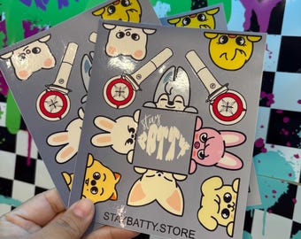 May include: A hand holding several gray stickers featuring cartoon animal designs, including bunnies, dogs, and cats. Each sticker has a unique design, with some featuring a red and white emblem. The text "stay batty.store" is visible.