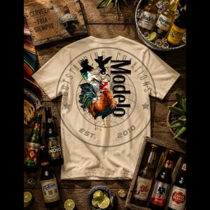 May include: Beige t-shirt with a circular graphic featuring a rooster, the word "Modelo", and the text "Outstanding Creations Est. 2010". The shirt is surrounded by beer bottles and limes.