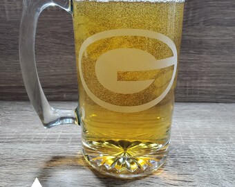 May include: A clear glass beer mug with a handle, filled with a light-colored beer. The mug has a green and white logo etched on the side, featuring a large "G" inside a circle.