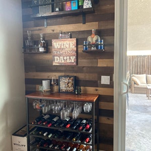 May include: A black metal wine rack with five shelves filled with bottles of wine. The rack is next to a wooden wall with shelves holding liquor bottles, glasses, and a sign that says "Wine Tasting is a Daily Thing".