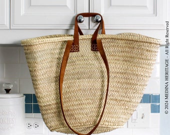May include: A large, natural woven basket with brown leather handles. The basket is hanging from a cabinet door.