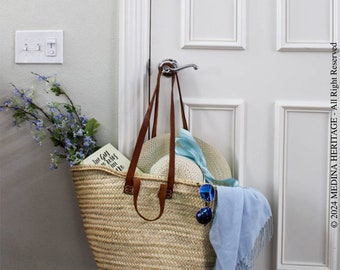 May include: A straw tote bag with brown leather handles hanging on a white door. The bag is filled with a straw hat, a blue scarf, and sunglasses. The text "I'm going to the beach" is visible on a book inside the bag.