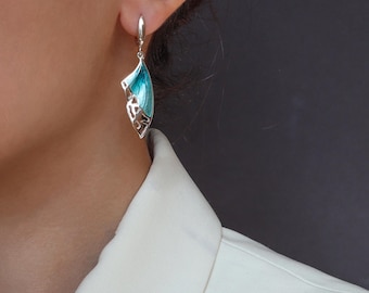 May include: A silver-toned drop earring featuring a curved, leaf-like shape with a vibrant turquoise blue enamel inlay. The metal structure has an openwork, filigree-style cutout pattern along the side, suspended from a simple silver-toned hook.