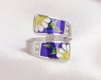 Könnte beinhalten: Ein Wickelring mit einer silbernen Ringschiene und zwei flachen, rechteckigen Enden. Jedes Ende zeigt eine farbenfrohe Emaille-Einlage mit einer weißen Blume mit gelbem Zentrum, grünen Blättern und einem tiefblauen Hintergrund.