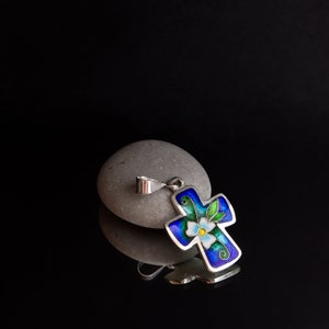 May include: A silver cross pendant with blue, green, and white enamel detailing. The cross features a stylized flower design.