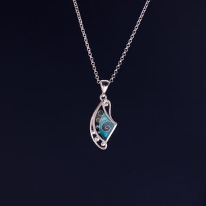 May include: A silver pendant necklace featuring a curved, fan-like shape with an openwork metal frame. The design includes a central teal enamel inlay with a spiral pattern, accented by small circular cutouts and silver spheres along the edge.