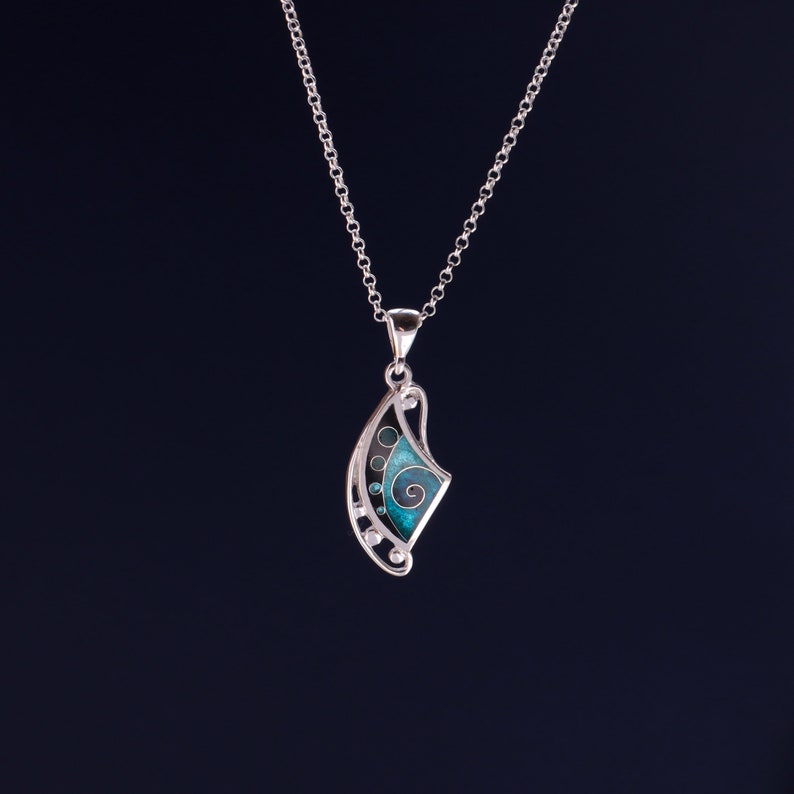 May include: A silver pendant necklace featuring a curved, fan-like shape with an openwork metal frame. The design includes a central teal enamel inlay with a spiral pattern, accented by small circular cutouts and silver spheres along the edge.