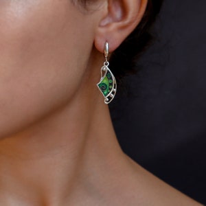 May include: A silver drop earring featuring a curved, crescent-shaped design with vibrant green enamel inlay. The piece is decorated with a black spiral pattern and small circular accents, suspended from a simple silver lever-back closure.
