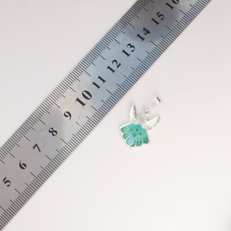 Op de afbeelding: Een zilveren engelhanger met groene emaille vleugels en een wit lichaam. De hanger is ongeveer 2 cm hoog en wordt getoond naast een liniaal.