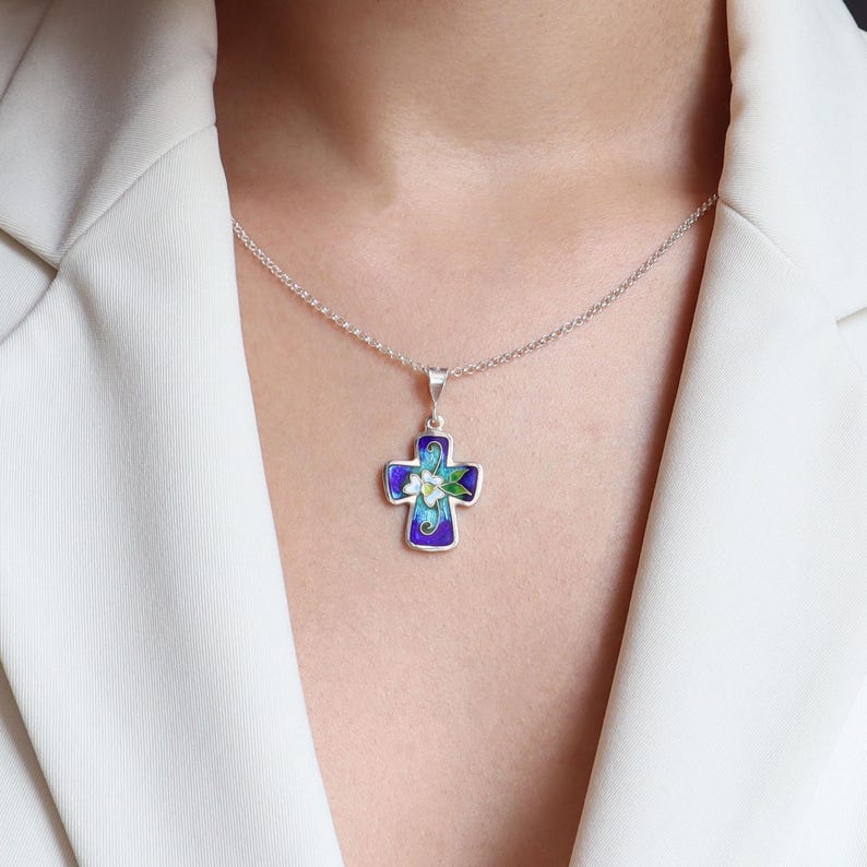 May include: A silver chain necklace with a blue and white enamel cross pendant featuring a floral design. The cross is decorated with white flowers and green leaves.