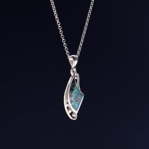 May include: A silver pendant necklace featuring a curved, asymmetrical shape with a dark background and a bright teal enamel inlay. The inlay displays a swirling spiral pattern and small circular accents. The pendant hangs from a silver chain.