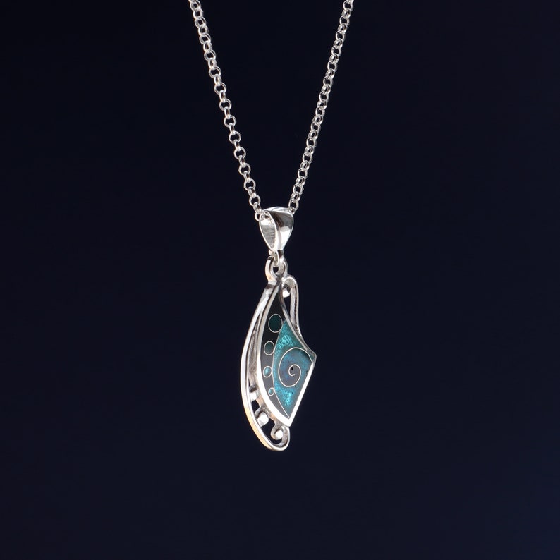 May include: A silver pendant necklace featuring a curved, asymmetrical shape with a dark background and a bright teal enamel inlay. The inlay displays a swirling spiral pattern and small circular accents. The pendant hangs from a silver chain.