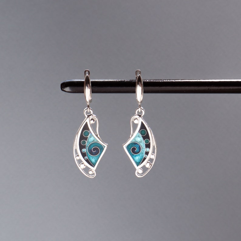 May include: A pair of silver drop earrings featuring a curved, fan-like shape with enamel inlay. The design includes a central dark blue spiral, surrounded by gradients of turquoise and teal, accented with small black circles and silver dots.