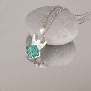 Op de afbeelding: Een zilveren ketting met een groene en witte emaille engel hanger. De engel heeft vleugels en een halo. De ketting ligt op een grijze steen.
