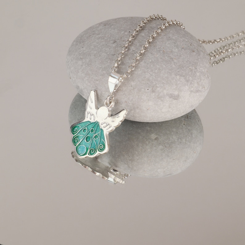 Op de afbeelding: Een zilveren ketting met een groene en witte emaille engel hanger. De engel heeft vleugels en een halo. De ketting ligt op een grijze steen.
