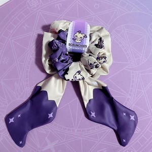 Op de afbeelding: Een crème- en paarse scrunchie set met een Mimikyu-ontwerp. De set bevat een lint met steraccenten. De verpakking luidt "SCRUNCHIES MIMIKYU".