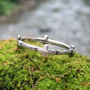 May include: A silver ring with a cross design. The ring is made of a thin band of metal with small crosses spaced evenly around the band. The ring is sitting on a bed of green moss.
