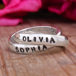 May include: Two silver rings intertwined, with the names "Olivia" and "Sophia" engraved on them. The rings are on a wooden surface.