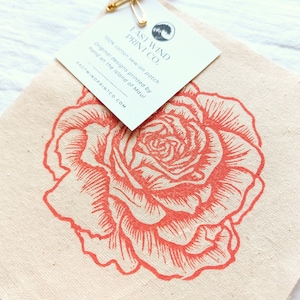 Puede incluir: Un parche de tela de color natural con una rosa de color coral impresa. El parche está etiquetado con "East Wind Print Co." y dice "100% algodón para coser. Diseños originales impresos a mano en la isla de Maui. Eastwindprintco.com."