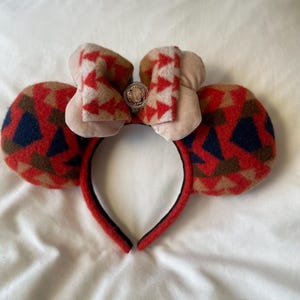 Può includere: Una fascia festiva con orecchie da topo. Le orecchie presentano un motivo di forme geometriche rosse, blu navy e beige. Un fiocco abbinato con un abbellimento argentato è in cima. La fascia è rossa con l'interno nero.