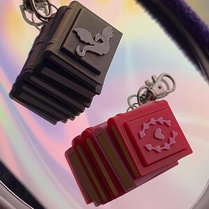 May include: Two book-shaped keychains with metal lobster clasps. One is black with a raised dragon emblem on the cover. The other is red with a raised heart surrounded by a circular pattern of small hearts on the cover. Both have layered page details.