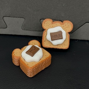 Op de afbeelding: Twee beervormige toastbedels met een getextureerde goudbruine korst, een glad wit roomcentrum en een klein bruin chocoladevierkantje met de tekst MILK in reliëf in het midden op de room. Dit zijn miniatuur replica's van voedsel.