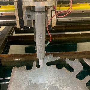 Puede incluir: Primer plano de una máquina de corte de metal con una boquilla de chorro de agua cortando una lámina de metal. La boquilla está etiquetada como "B.I.A.M."