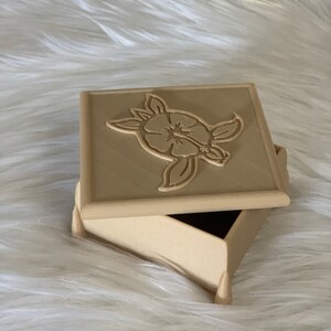 May include: A beige wooden box with a hinged lid. The lid features a carved design of a turtle with a flower on its back.