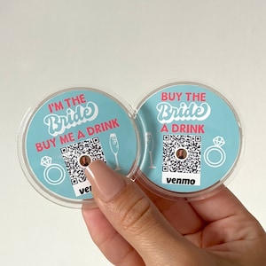 Könnte beinhalten: Zwei runde, blaue und weiße Aufkleber mit einem QR-Code und dem Text "I'm the Bride Buy me a drink" und "Buy the Bride a drink" mit einem Ring und einem Champagnerglas.