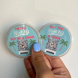 Könnte beinhalten: Zwei runde, blaue und weiße Papieruntersetzer mit einem rosa und weißen QR-Code. Der Text auf den Untersetzern lautet "I'm the Bride Buy Me a Drink" und "Buy the Bride a Drink".