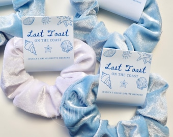 May include: Four blue and white velvet scrunchies with a white tag attached to each. The tag has a blue and white illustration of seashells and a starfish. The text on the tag reads "Last Toast on the Coast Jessica's Bachelorette Weekend".