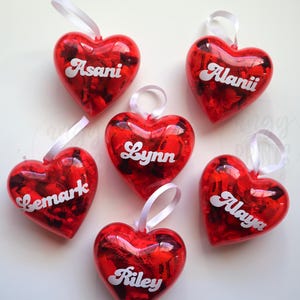 May include: Six red heart-shaped ornaments with white ribbon loops. Each heart has a different name in white script: Asani, Alanii, Lemark, Lynn, Alaya, and Riley. The hearts have a shiny, reflective surface.
