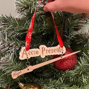 May include: Wooden Christmas ornament with the words "Accio Presents" and a wand design. The ornament is hanging on a Christmas tree with a red ribbon and a red glitter ball ornament. The name "Alayna" is also on the wand.
