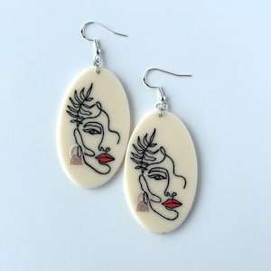 May include: A pair of cream-colored oval earrings with a black line drawing of a face with a leaf on the side. The face has red lips and a small pink and brown design on the side.