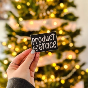 May include: A black sticker with white text that reads "Product of Grace" held in a hand against a blurry background of a Christmas tree with white lights.