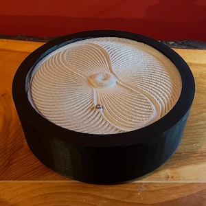 Könnte beinhalten: Ein schwarzer, runder Zen-Garten mit einem hellbeige Sandmuster. Der Sand ist in einem Spiralmuster geharkt, mit einer kleinen silbernen Kugel in der Mitte. Der Garten steht auf einer Holzoberfläche.