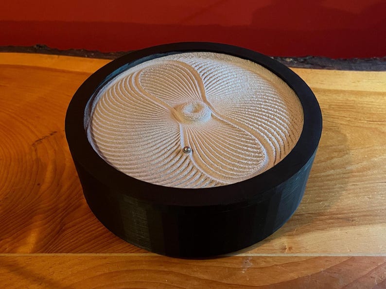 Puede incluir: Un jard&iacute;n zen redondo y negro con un dise&ntilde;o de arena beige claro. La arena est&aacute; rastrillada en un patr&oacute;n en espiral con una peque&ntilde;a bola plateada en el centro. El jard&iacute;n est&aacute; sobre una superficie de madera.
