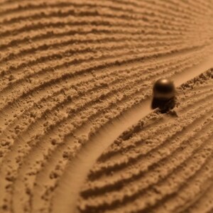 Peut inclure: Gros plan sur un jardin zen avec une boule roulant dans le sable. Le sable est brun clair et présente une surface texturée avec des lignes courbes. La boule est sombre et métallique.