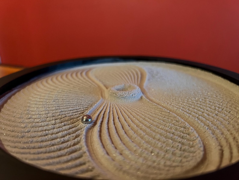 Puede incluir: Primer plano de una pieza de arte cin&eacute;tico de arena. La arena es de color beige claro con un patr&oacute;n de l&iacute;neas curvas que irradian desde un punto central. Una peque&ntilde;a bola de metal plateado descansa sobre la arena, creando el dise&ntilde;o. La pieza est&aacute; en un marco circular negro.