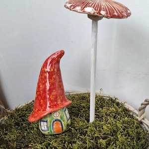 Könnte beinhalten: Ein Keramik-Pilzhaus mit einem hohen, geschwungenen roten Dach mit gelben Sprenkeln, einem hellgrünen Sockel und einer kleinen orangefarbenen Tür. Daneben steht ein langer, dünner weißer Stiel mit einem breiten, flachen, gerippten roten Pilzhut.