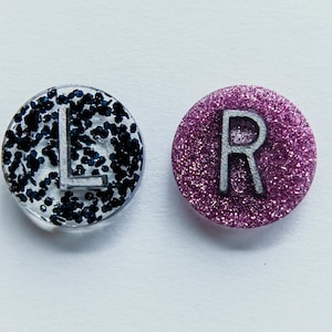 May include: Two round, glittery resin pieces, one black and silver, the other purple. The black piece has the letter "L" in silver, and the purple piece has the letter "R" in silver. Both are on a white background.