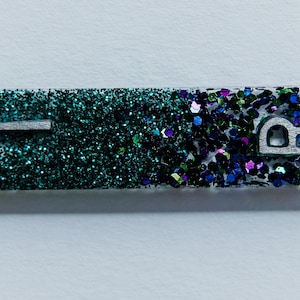 Könnte beinhalten: Ein rechteckiges Stück mit teal- und lilafarbenem Glitzer mit den Buchstaben "L" und "R" in Weiß.