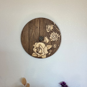 Op de afbeelding: Een ronde houten wanddecoratie met een houtnerfpatroon en een ontwerp van witte rozen die in het hout zijn gebrand.