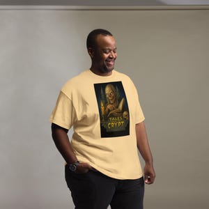 May include: A light tan t-shirt featuring a graphic from "Tales from the Crypt." The graphic shows a skeletal figure with a candle and a skull. The shirt is a relaxed fit and has short sleeves.
