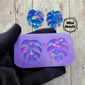 May include: Purple silicone mold with two monstera leaf shapes. Two iridescent blue monstera leaf earrings are shown next to the mold.