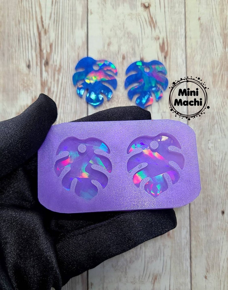 May include: Purple silicone mold with two monstera leaf shapes. Two iridescent blue monstera leaf earrings are shown next to the mold.