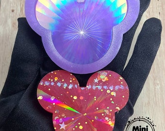 May include: Two resin Mickey Mouse-shaped items. One is a light purple color with a holographic design. The other is red with glitter and star accents. The items are displayed on a black surface. The Mini Machi logo is visible.