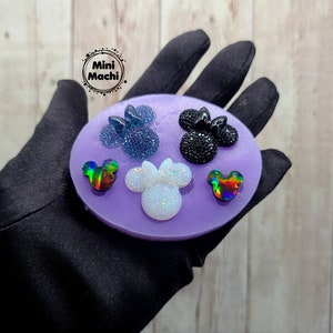 May include: A purple silicone mold with five different mouse ear shapes. The shapes are filled with colorful glitter resin. The mold is on a black surface.