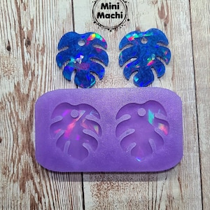 May include: Two iridescent blue resin monstera leaf earrings with a hole in the center. The earrings are next to a purple silicone mold with two monstera leaf shapes.