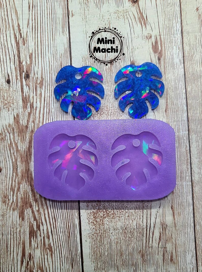 May include: Two iridescent blue resin monstera leaf earrings with a hole in the center. The earrings are next to a purple silicone mold with two monstera leaf shapes.